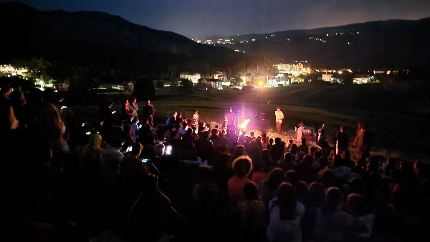 Ragazzi del camp seduti intorno a un falò la sera, con le luci della valle sullo sfondo