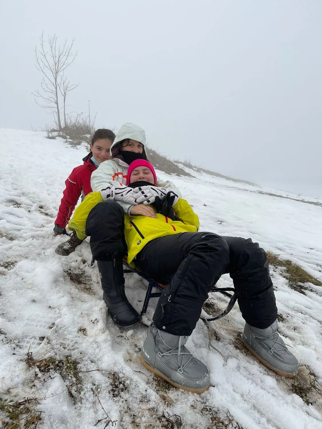 Tre ragazzi del Camp sulla Neve seduti uno dietro l’altro sullo stesso slittino, abbracciati e sorridenti, su un pendio innevato in una giornata di nebbia